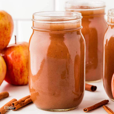 A glass jar filled with rich, brown apple butter, its smooth texture visible through the jar. The lid is tightly sealed, and a small wooden spoon rests beside it, ready to spread the sweet, spiced spread. The background features a rustic wooden table with fresh apples and a sprinkling of cinnamon sticks, evoking cozy, homemade vibes.