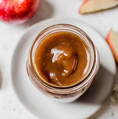 A glass jar filled with rich, brown apple butter, its smooth texture visible through the jar. The lid is tightly sealed, and a small wooden spoon rests beside it, ready to spread the sweet, spiced spread. The background features a rustic wooden table with fresh apples and a sprinkling of cinnamon sticks, evoking cozy, homemade vibes.
